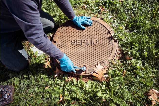 A person wearing blue gloves is kneeling on grass and using a wrench to open a round, rusty metal septic tank lid with "SEPTIC" embossed on it, TO PROVIDE septic system service central valley