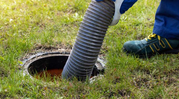 A close-up, overhead view of a green and black septic tank riser opening with a person's gloved hand holding a large, ribbed green and yellow pumping hose inserted into the murky liquid inside.