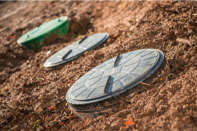 Three round plastic septic tank lids (one green, two grey) are visible above ground, set in damp, reddish-brown soil, possibly indicating areas over the tank or drain field distribution.