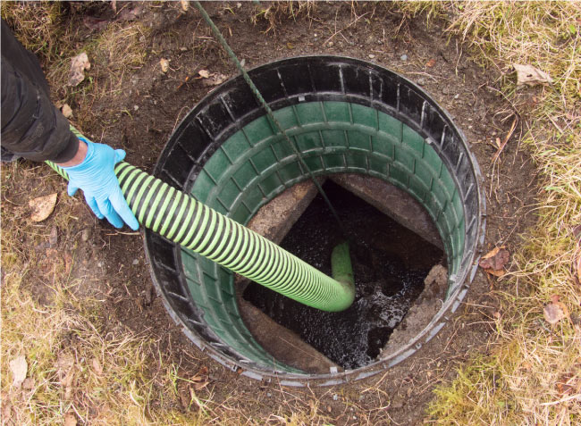 A close-up, overhead view of a green and black septic tank riser opening with a person's gloved hand holding a large, ribbed green and yellow pumping hose inserted into the murky liquid inside.