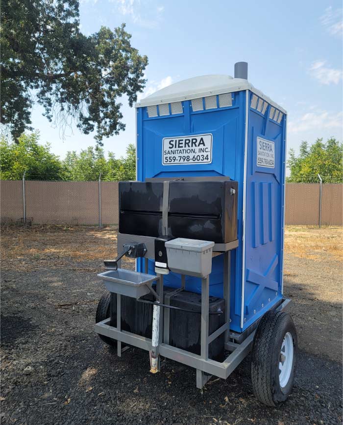 Blue towable porta potty rental on a single-axle trailer with an integrated handwashing sink station in a dry field.
