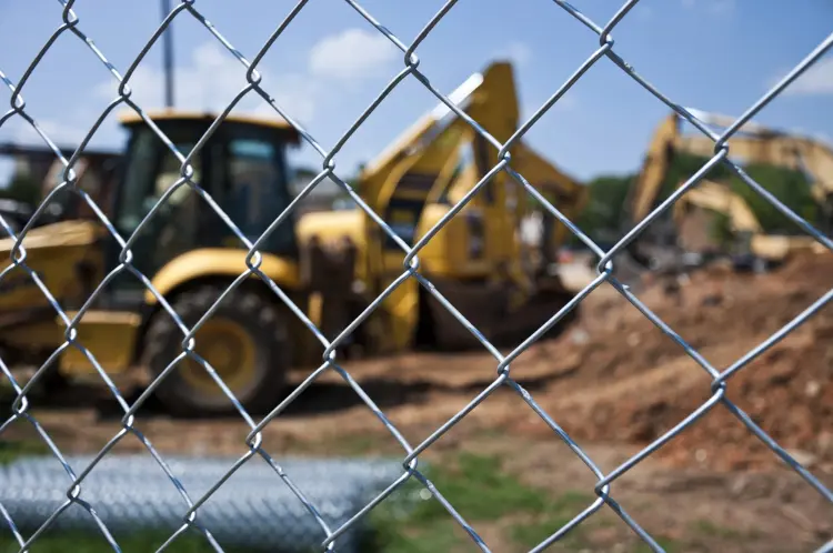 Close-up view of a silver chain-link temporary fence securing a construction site with heavy yellow earth-moving machinery blurred in the background.