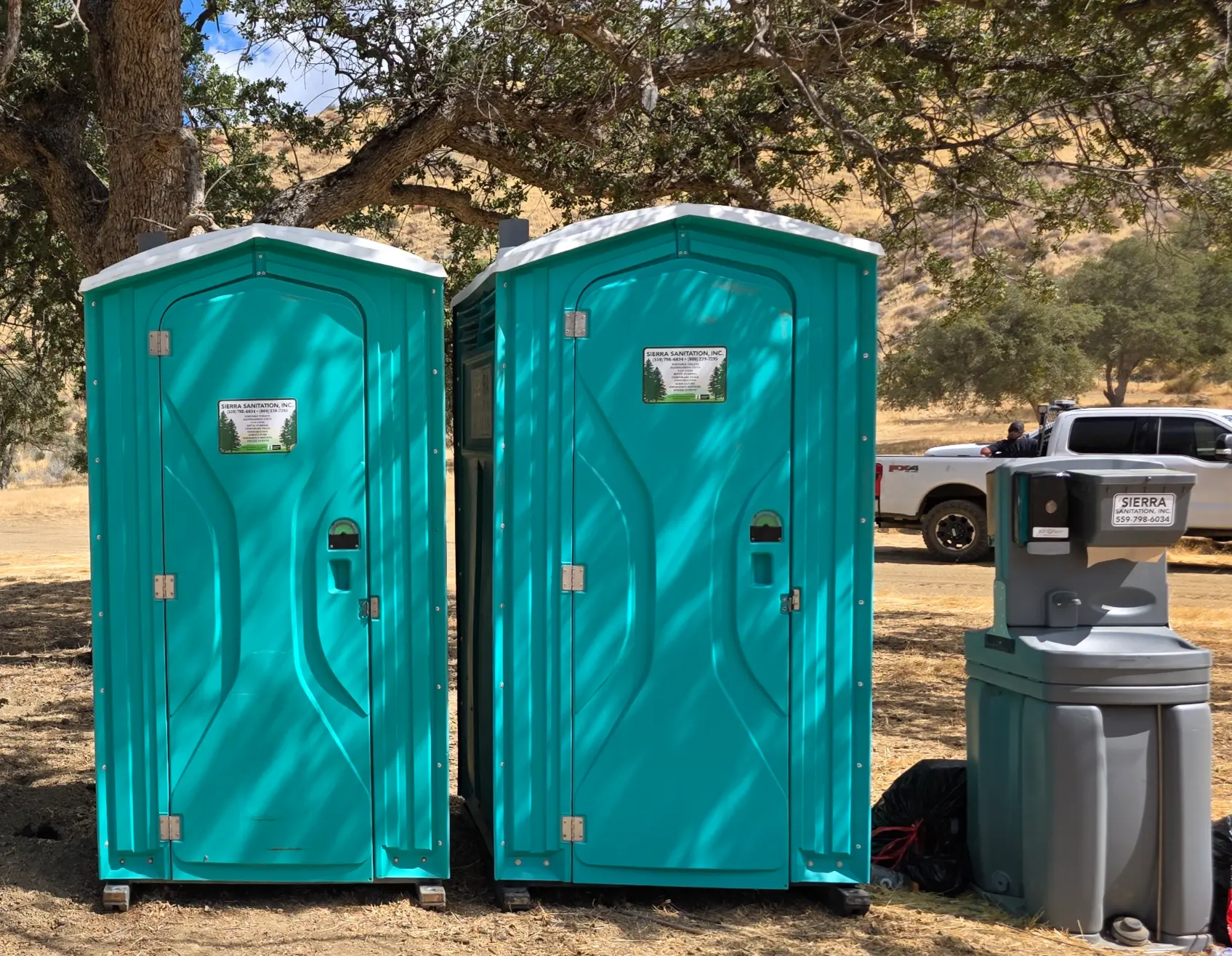 Two teal portable toilet units and a grey outdoor handwashing station from Sierra Sanitation placed outdoors on a job site near a white pickup truck.